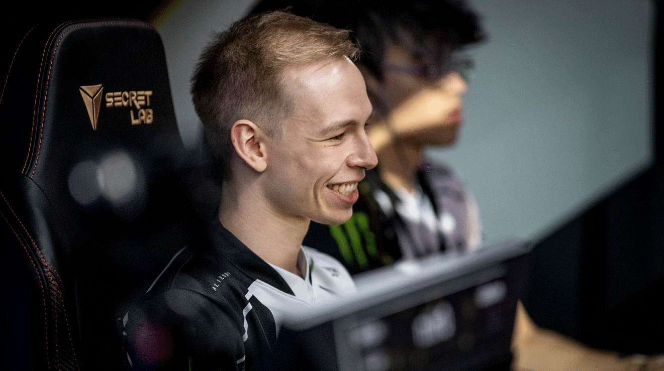 EliGE in front of his monitor, smiling as he jokes with his team mates who are off frame