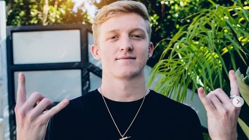 Tfue stands outside, he faces the camera with hands raised in the hang loose sign. Green trees and blue sky appear behind him.