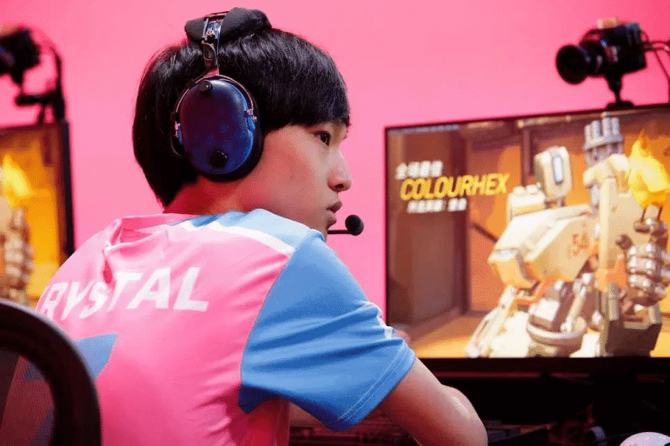 Overwatch League player Cai "Krystal" Shilong in front of a PC monitor showing Overwatch hero Bastion