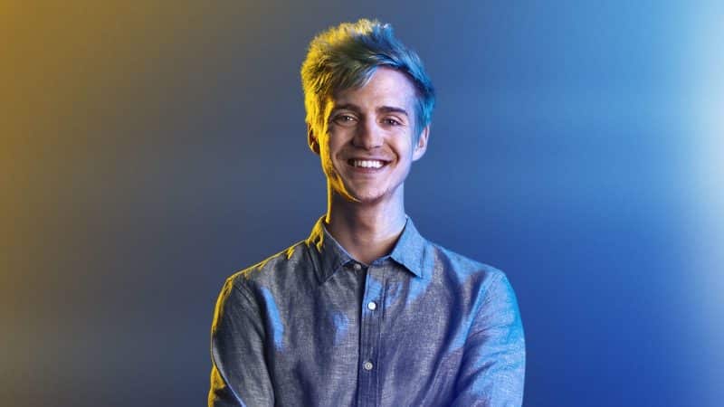 Tyler "Ninja" Blevins stsmiles for the camera under blue and yellow tinted lights. He is wearing a blue, collared shirt with his top button undone.