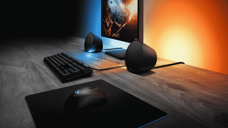 A computer setup is shown with a monitor, keyboard, mouse and the Logitech G560 speakers with blue and yellow lights behind it all
