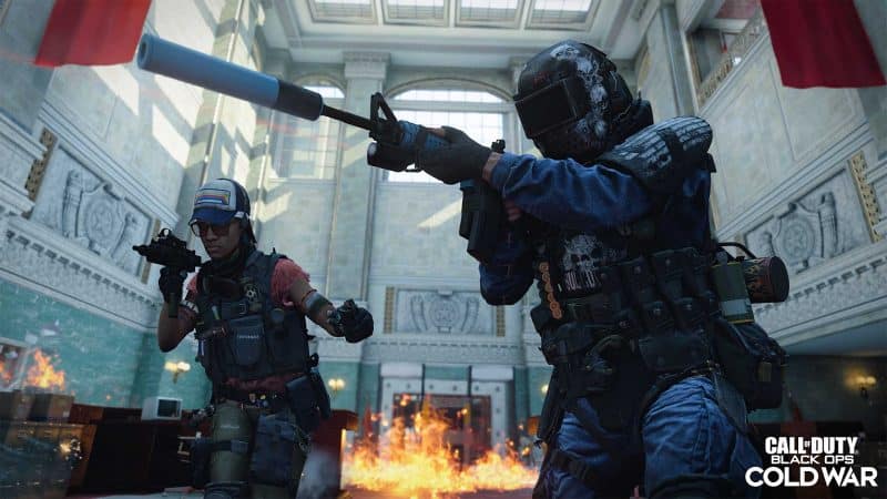 Two military operators clear a large office room with fire overtaking the floor and red flags on the high ceiling and the words "Call of Duty Black Ops Cold War" in the bottom left corner