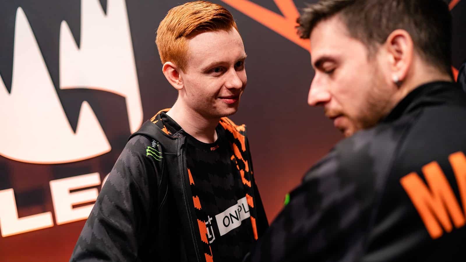 Pro LoL player Daniel “Dan” Hockley smiling at a Fnatic teammate with the LEC crown logo on the wall behind him