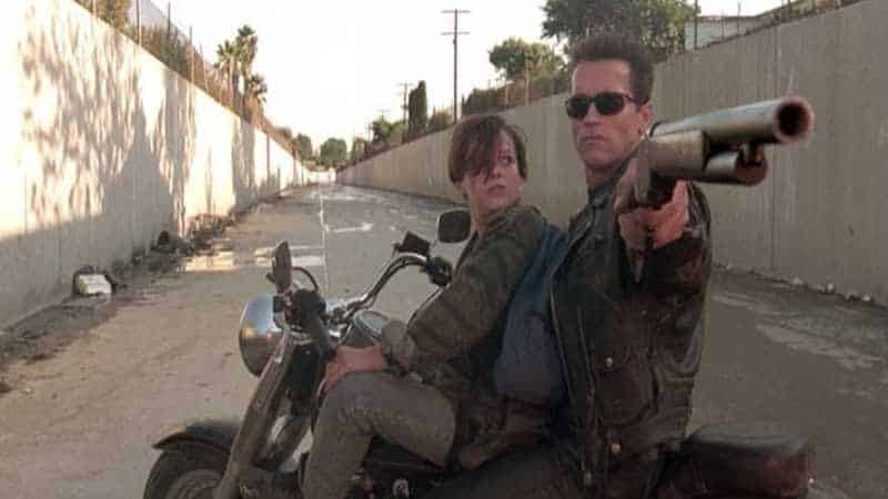 Arnold Schwarzenegger sits on a motorbike alongside a young John Connor, pointing his shotgun at the camera in Terminator 2: Judgement Day