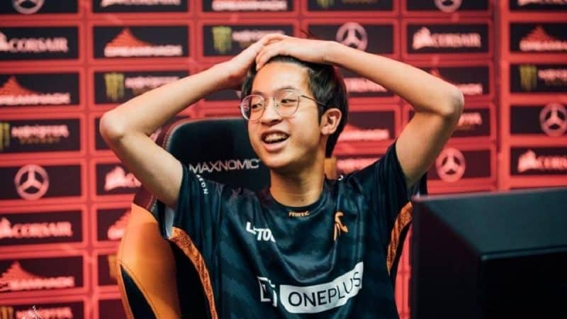 Pro Dota 2 player Nuengnara “23savage” Teeramahanon puts his hands on his head with an expression of disbelief on his face. He wears a Fnatic jersey in front of a PC monitor with a background filled with brand logos behind him.