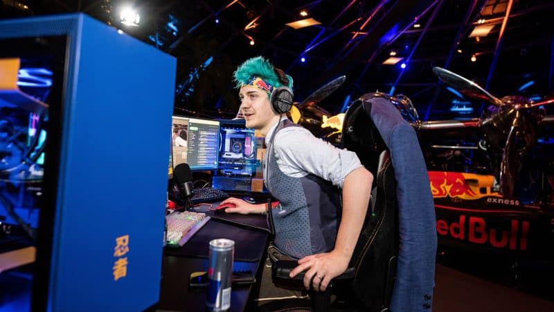 Pro gamer and streamer Tyler "Ninja" Blevins starts to stand up from a gaming chair in a stadium filled with lights and in front of a gaming PC with a Red Bull banner behind him