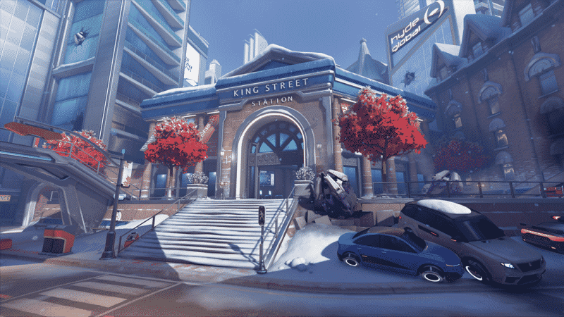 A screenshot from a new Overwatch map depicting Toronto's King Street Station in the winter time. A bit of snow lines the sidewalks, cars and buildings and three red trees stand tall outside of the main station building.