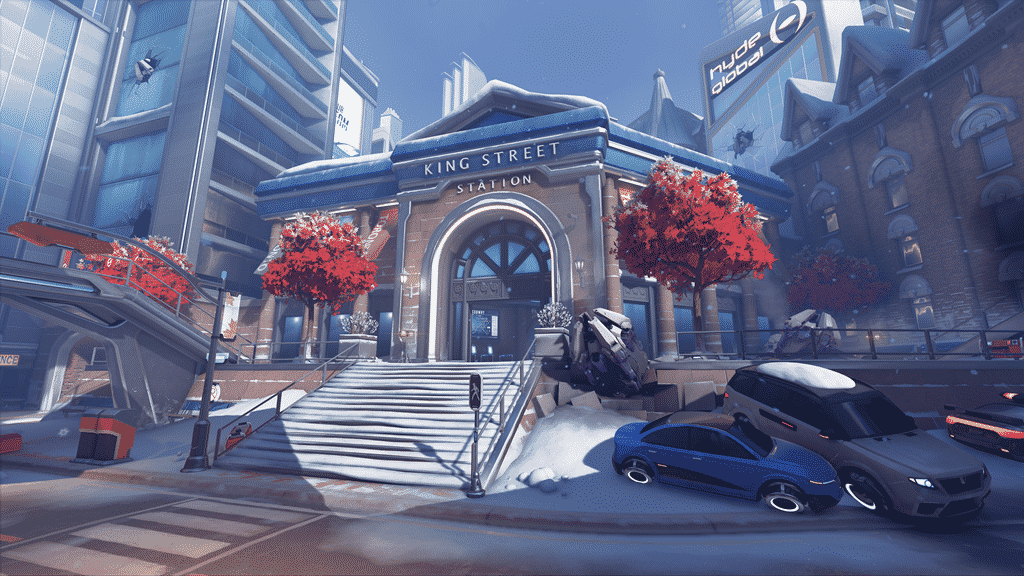 A screenshot from a new Overwatch map depicting Toronto's King Street Station in the winter time. A bit of snow lines the sidewalks, cars and buildings and three red trees stand tall outside of the main station building.
