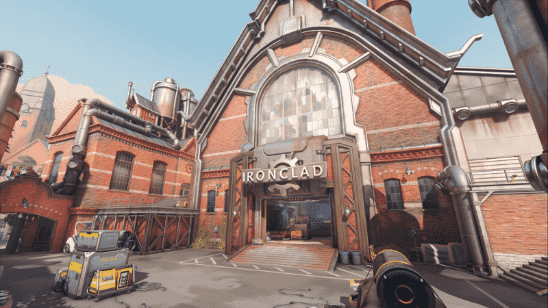 A screenshot from a new Overwatch map showing a large, brick industrial building with a sign out front with the word "Ironclad" with a gear next to it. Some machinery is scattered in front and inside.