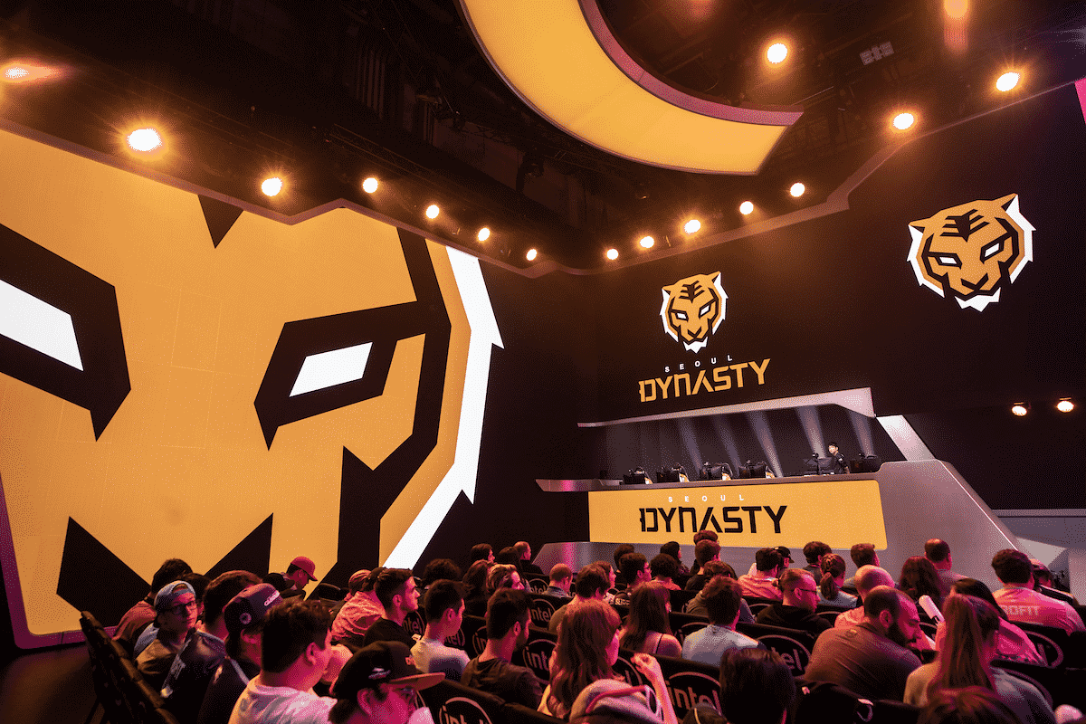 The Overwatch League arena is lit up by orange lights and boasts a large crowd in the stands. The stage contains a number of large monitors, each of them showing off the Seoul Dynasty's tiger logo.