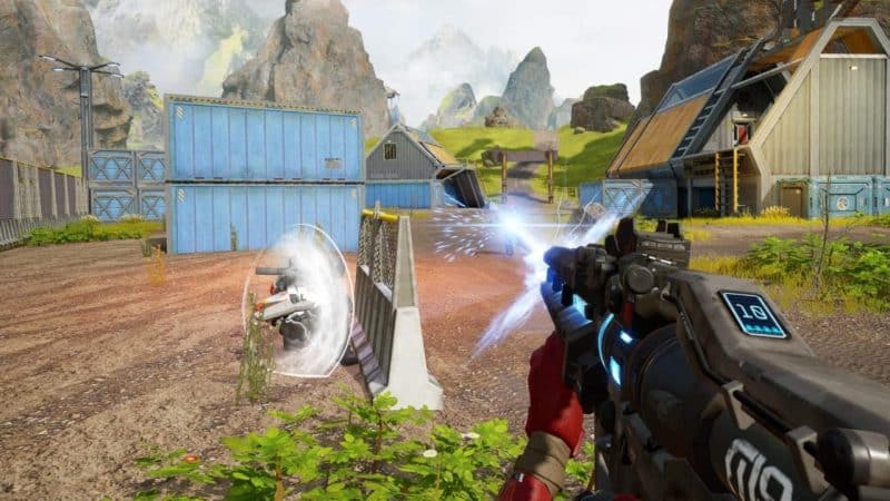An ingame screenshot from Apex Legends Mobile shows a player firing their weapon through sea containers and barricades