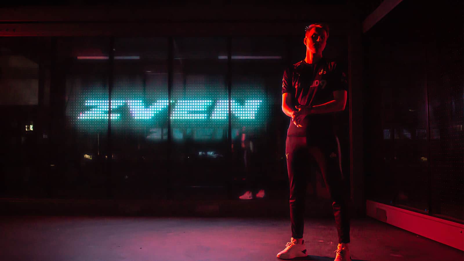 Pro LoL player Jesper “Zven” Svenningsen stands in a dimly lit room with his tag "Zven" in blue lights behind him.