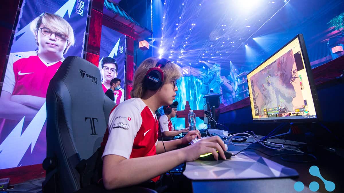 Kenny "Xepher" Deo and Karl "Karl" Baldovino sit side by side as they compete on the stage, the screens behind them show full screen photos of them in their T1 team jerseys