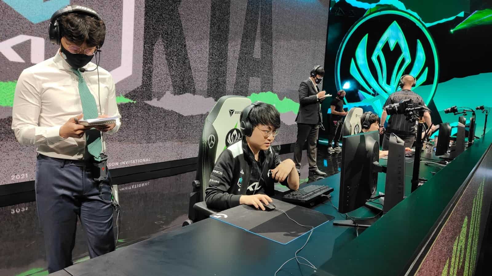 DAMWON KIA's mid laner Heo “Showmaker” Su sits on the MSI 2021 stage with his teammates at their PCs and his coach standing next to him.